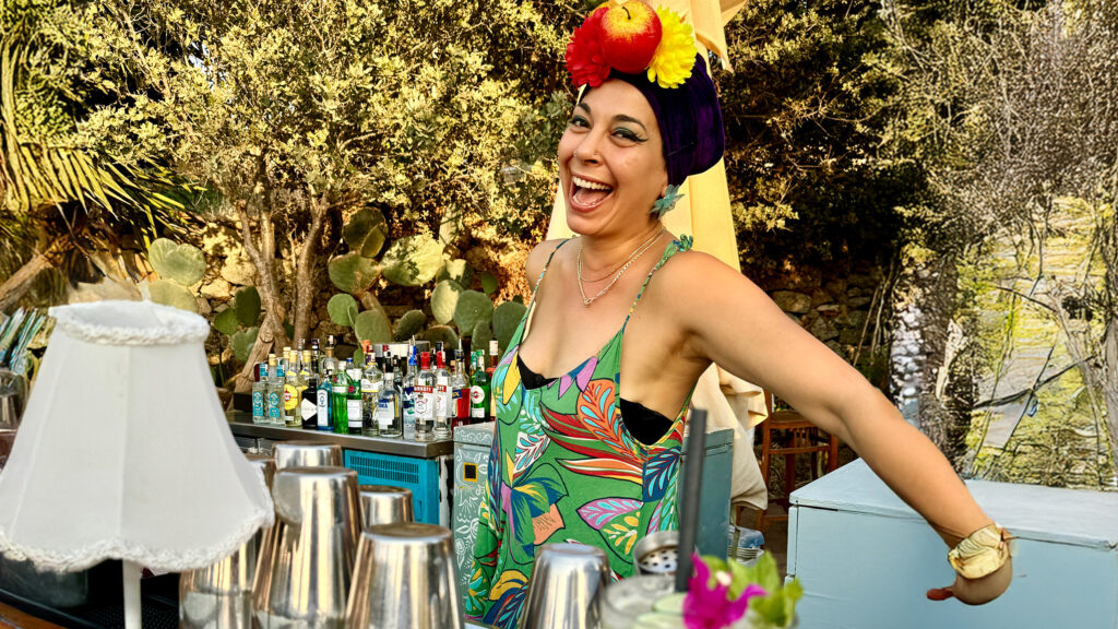 Fröhliche Bartenderin mit Obst-Turban und buntem Kleid hinter einer Open-Air-Bar voller Cocktailzutaten