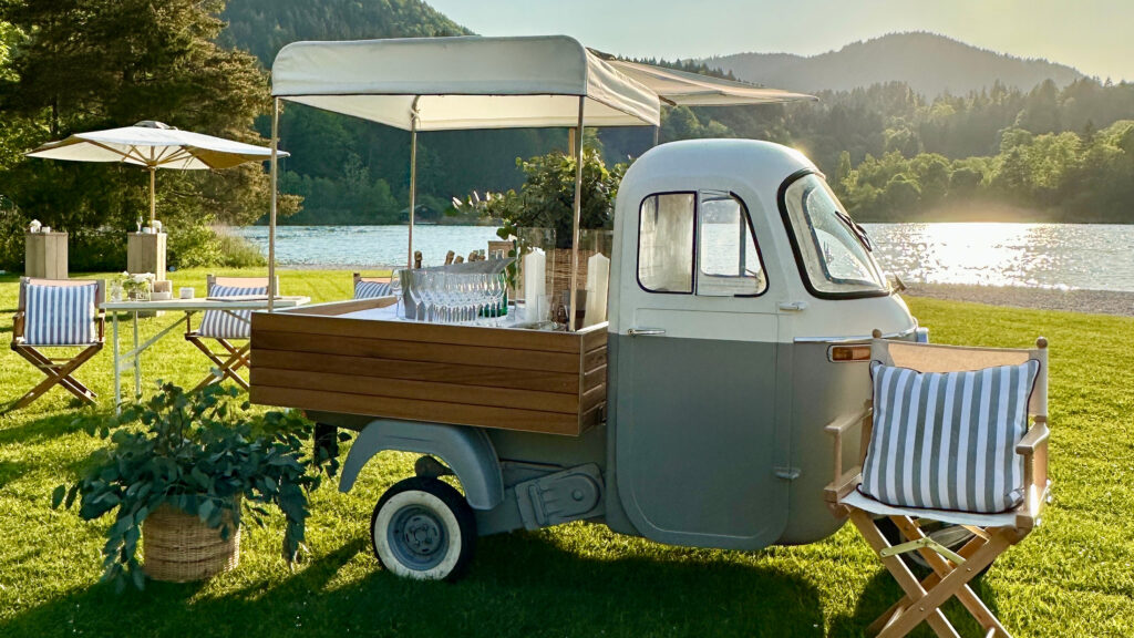 Kleine mobile Bar in einer Ape mit Gläsern und Dekoration, aufgestellt auf einer Wiese am See mit Liegestühlen und Sonnenschirmen